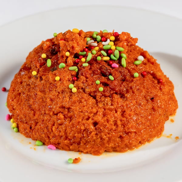 A vibrant orange Nepalese dessert, possibly a halwa, topped with colorful sprinkles on a white plate.