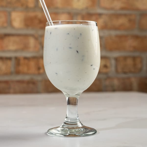 A glass of white Nepali lassi with dark specks and a straw, against a brick wall.