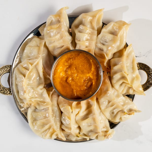A silver platter of Nepalese momo dumplings surrounds a small bowl of orange dipping sauce.