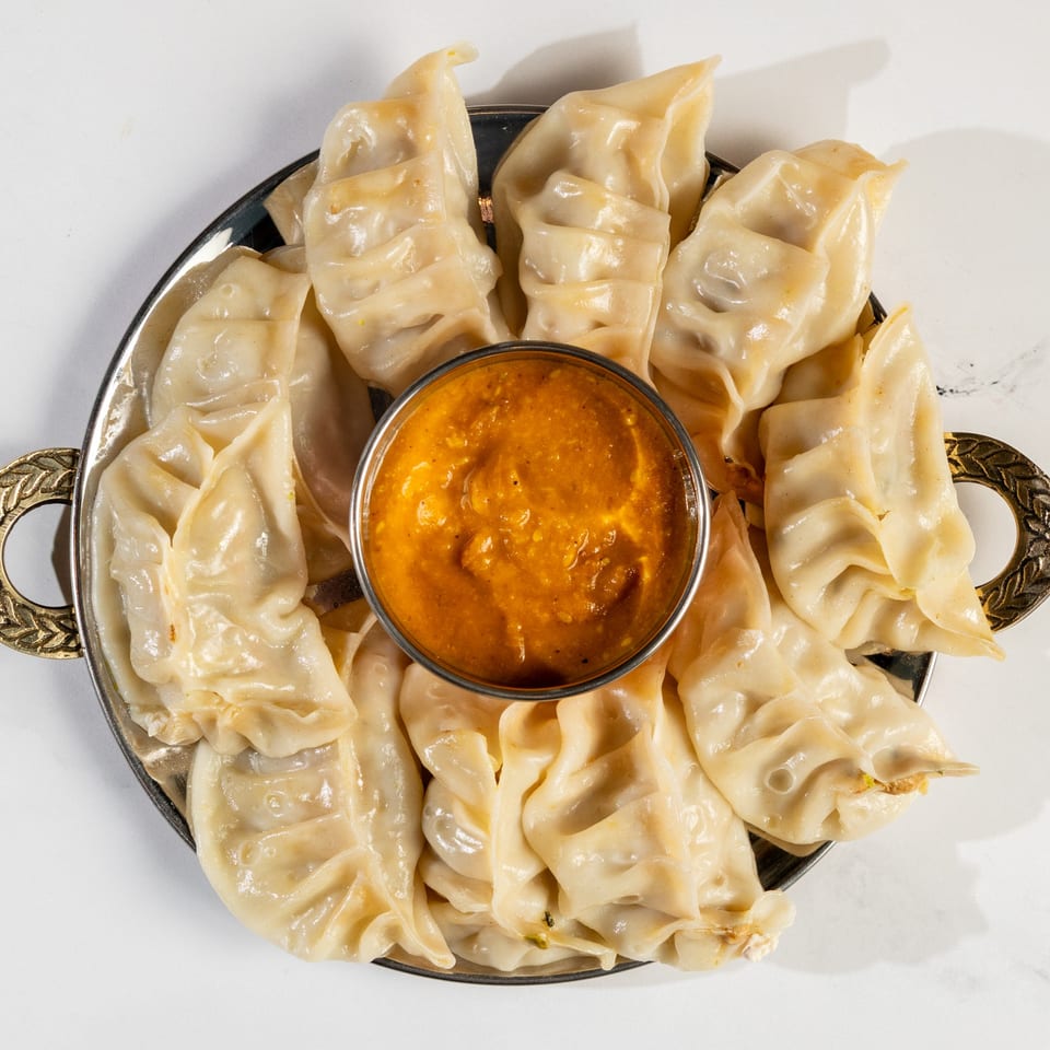 A silver platter of Nepalese momo dumplings surrounds a small bowl of orange dipping sauce.