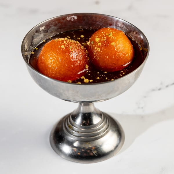 Two golden-brown gulab jamun in a silver bowl, soaked in dark syrup and topped with crushed nuts.